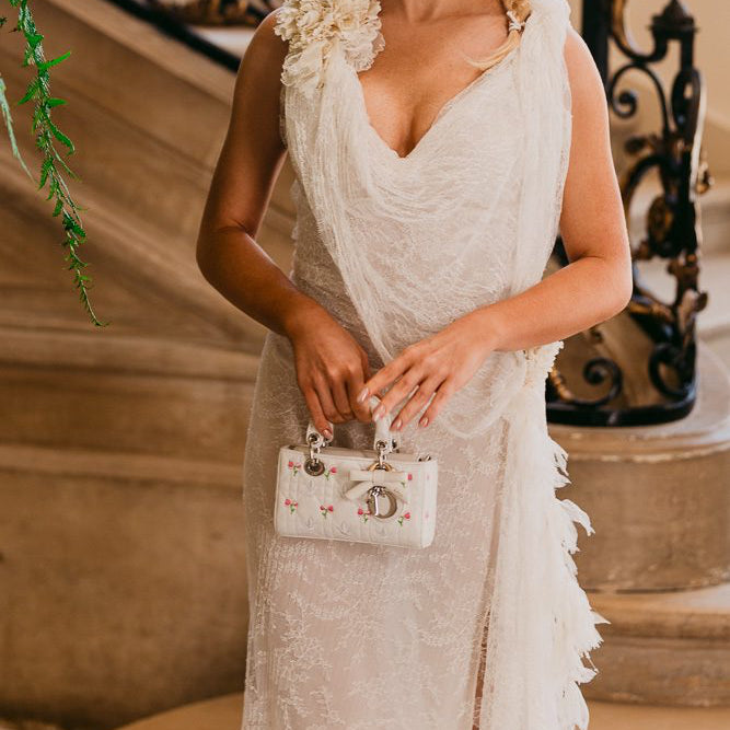 A woman wearing a sleeveless white lace dress with a draped neckline and floral applique stands on an indoor staircase, holding a small white handbag with pink floral embroidery and silver hardware.