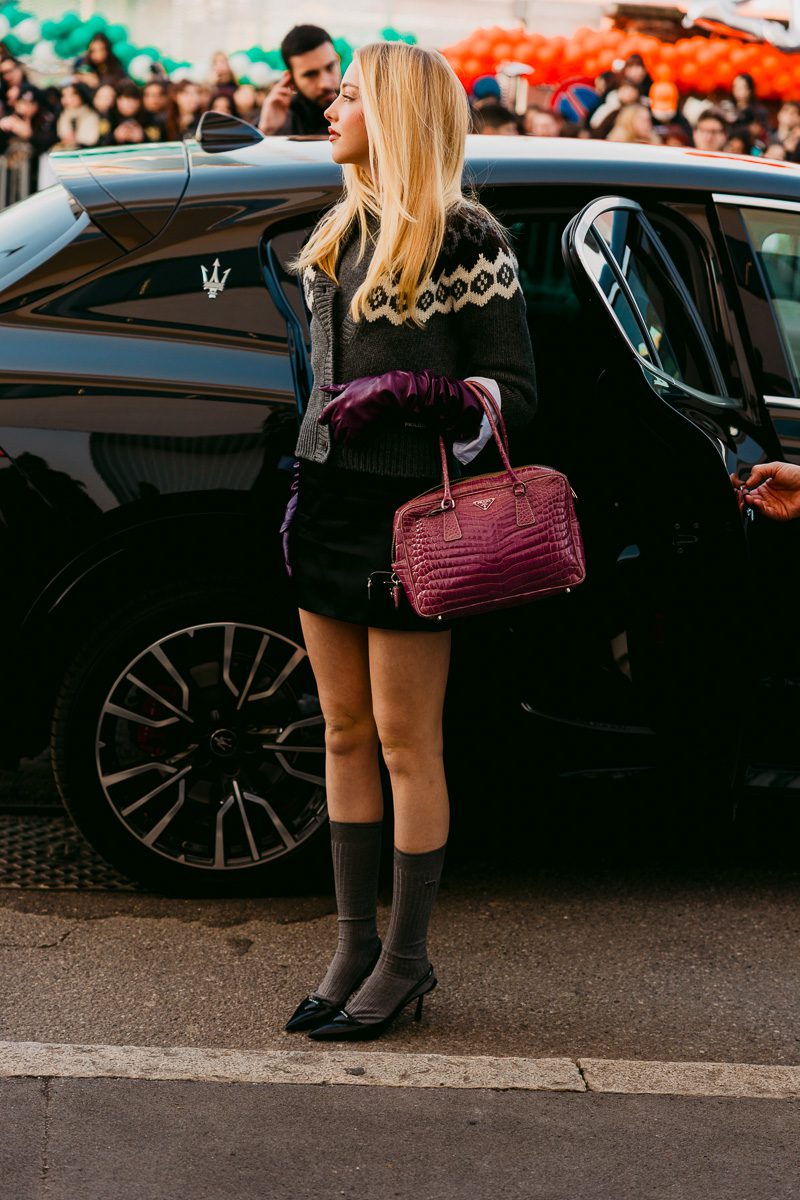 A woman stands beside a black car, wearing a dark knit sweater with a cream geometric pattern, a black skirt, gray knee-high socks, and black heels, while carrying a structured magenta handbag.