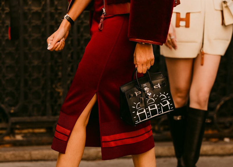 A fashion street-style shot featuring a model wearing a deep burgundy velvet midi skirt with a side slit, matching velvet jacket, and dark burgundy pointed-toe ankle boots. The model is holding a black handbag with white monogram lettering, set against a blurred urban background.