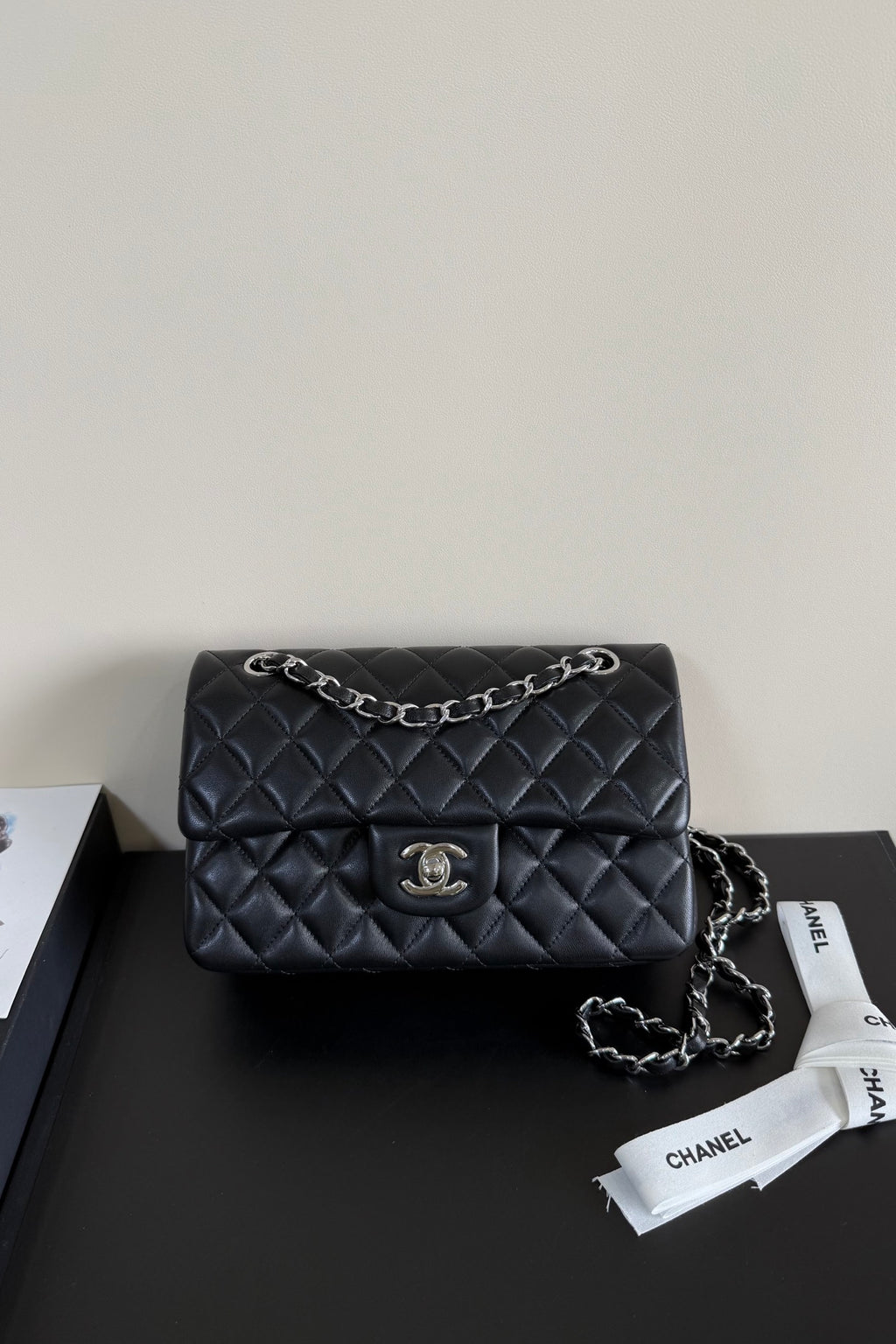 A black quilted Chanel flap shoulder bag with silver-tone chain strap and CC turn-lock closure, displayed on a dark surface with a Chanel dust bag ribbon nearby.
