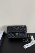 A black quilted Chanel flap shoulder bag with silver-tone chain strap and CC turn-lock closure, displayed on a dark surface with a Chanel dust bag ribbon nearby.