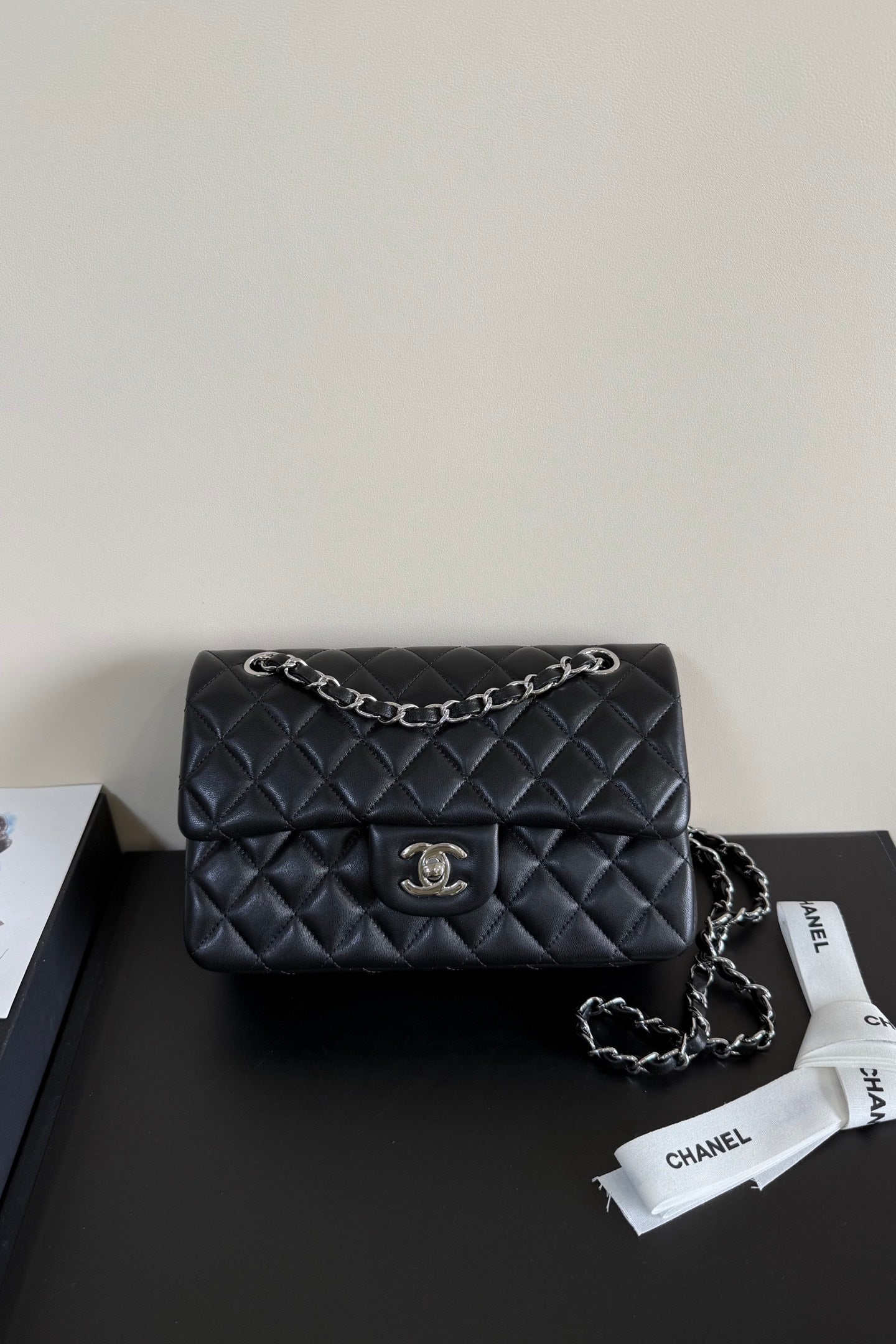 A black quilted Chanel flap shoulder bag with silver-tone chain strap and CC turn-lock closure, displayed on a dark surface with a Chanel dust bag ribbon nearby.