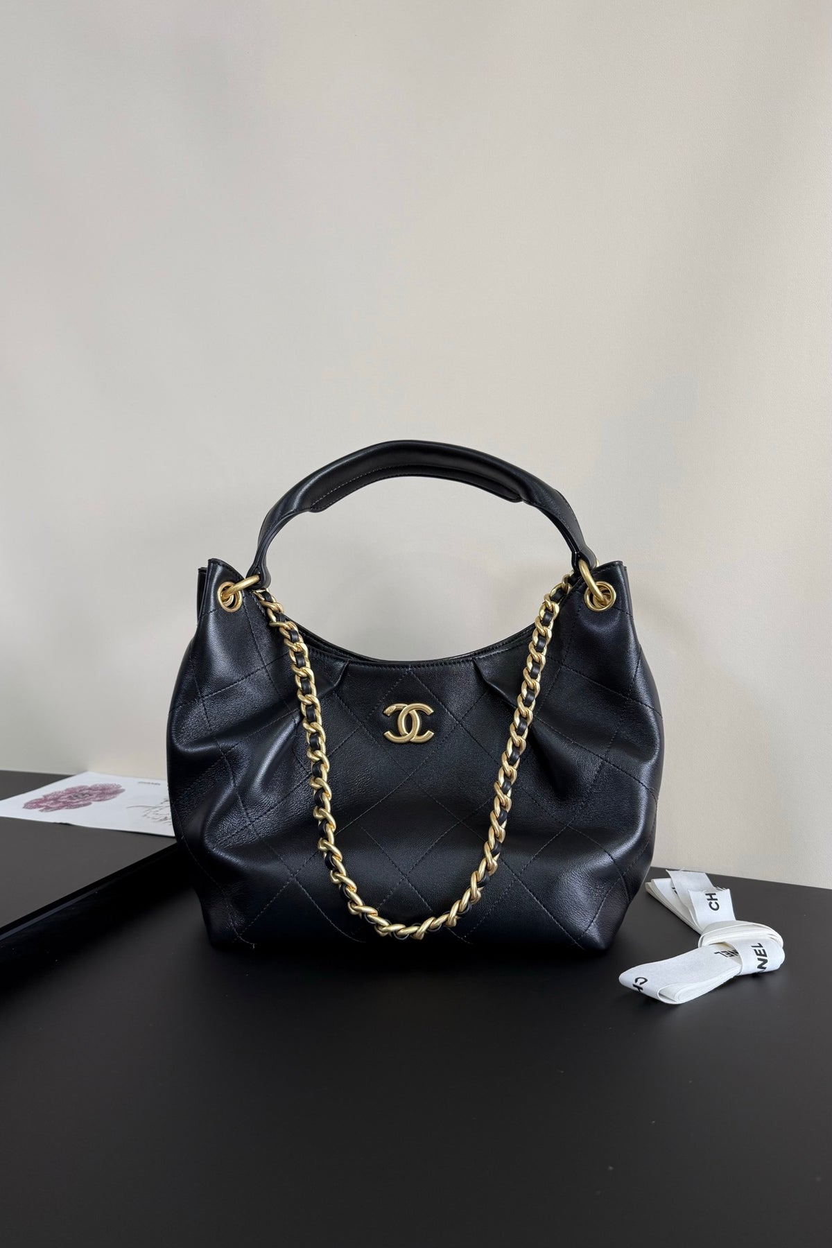 A black quilted leather handbag with gold-tone hardware, featuring a top handle, a gold chain strap, and a prominent interlocking CC logo on the front, displayed on a dark surface against a light wall.