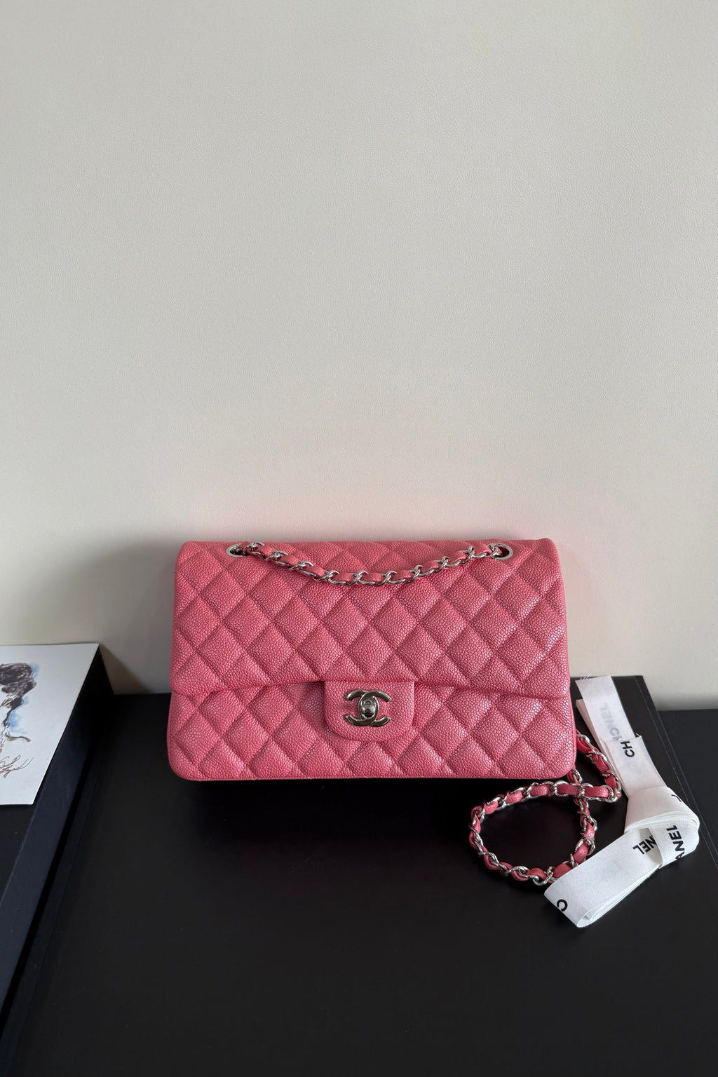 A pink quilted leather handbag with a silver-tone chain strap and a silver interlocking CC logo clasp, resting on a black surface next to a white branded ribbon.