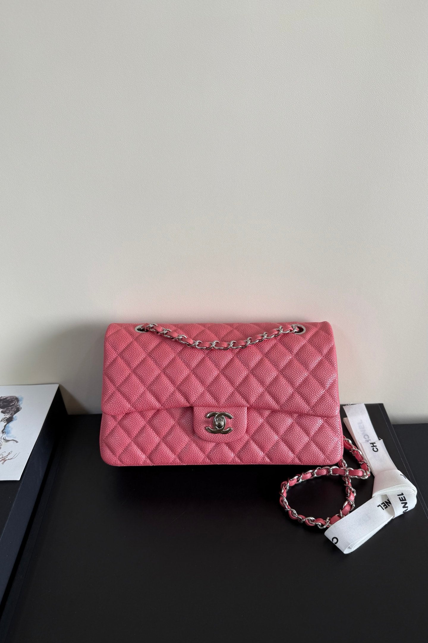 A pink quilted leather handbag with a silver-tone chain strap and a silver interlocking CC logo clasp, resting on a black surface next to a white branded ribbon.