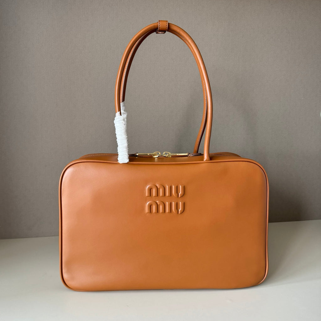 A Miu Miu tan leather handbag with a structured rectangular shape, double top handles, and a white protective paper sleeve wrapped around one handle. The embossed Miu Miu logo is visible on the front, set against a neutral gray background.