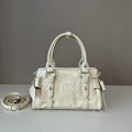 A cream-colored Miu Miu leather handbag with silver-tone hardware, displayed on a light gray surface against a dark gray background, with its matching shoulder strap coiled to the left.