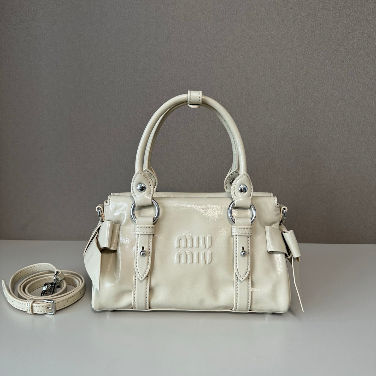 A cream-colored Miu Miu leather handbag with silver-tone hardware, displayed on a light gray surface against a dark gray background, with its matching shoulder strap coiled to the left.