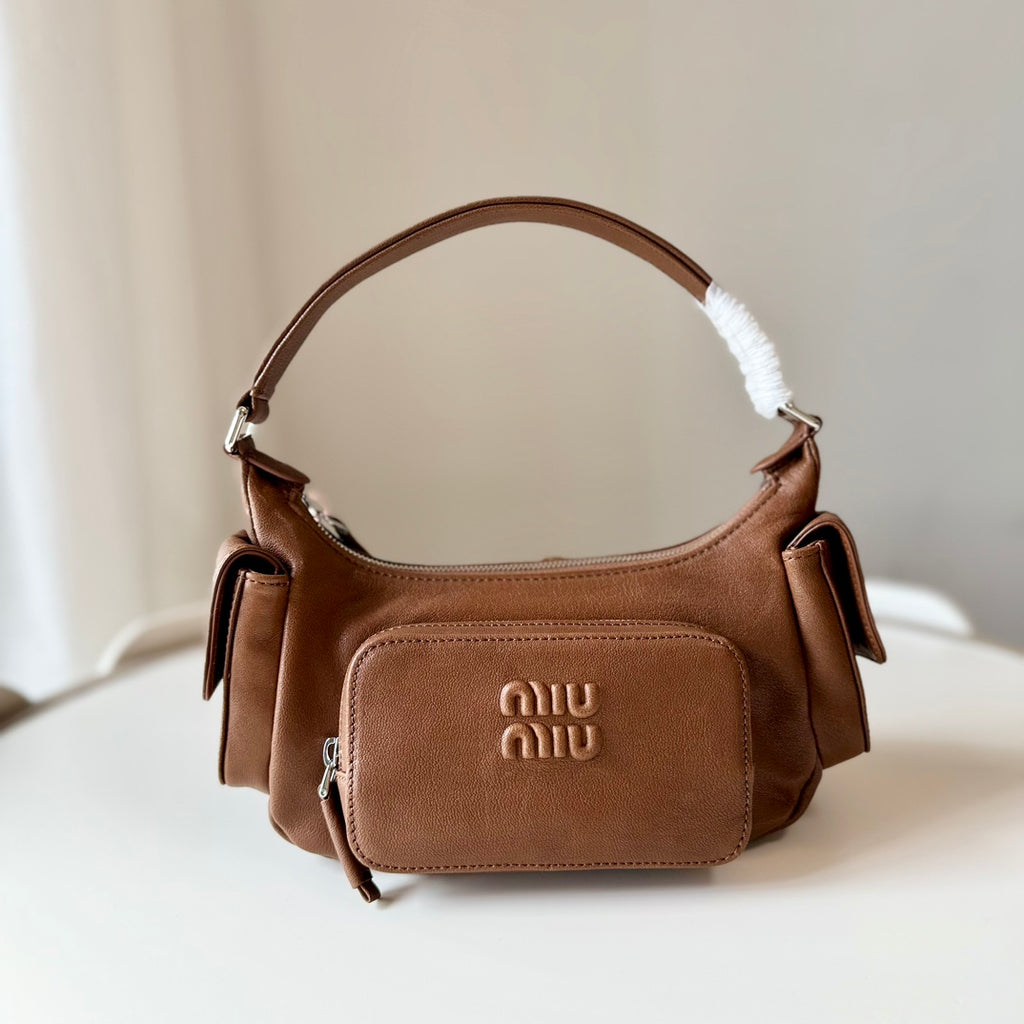 A brown leather shoulder bag with a front zip pocket embossed with 'miu miu' and a white faux-fur trim on the handle, resting on a white surface against a soft beige background.