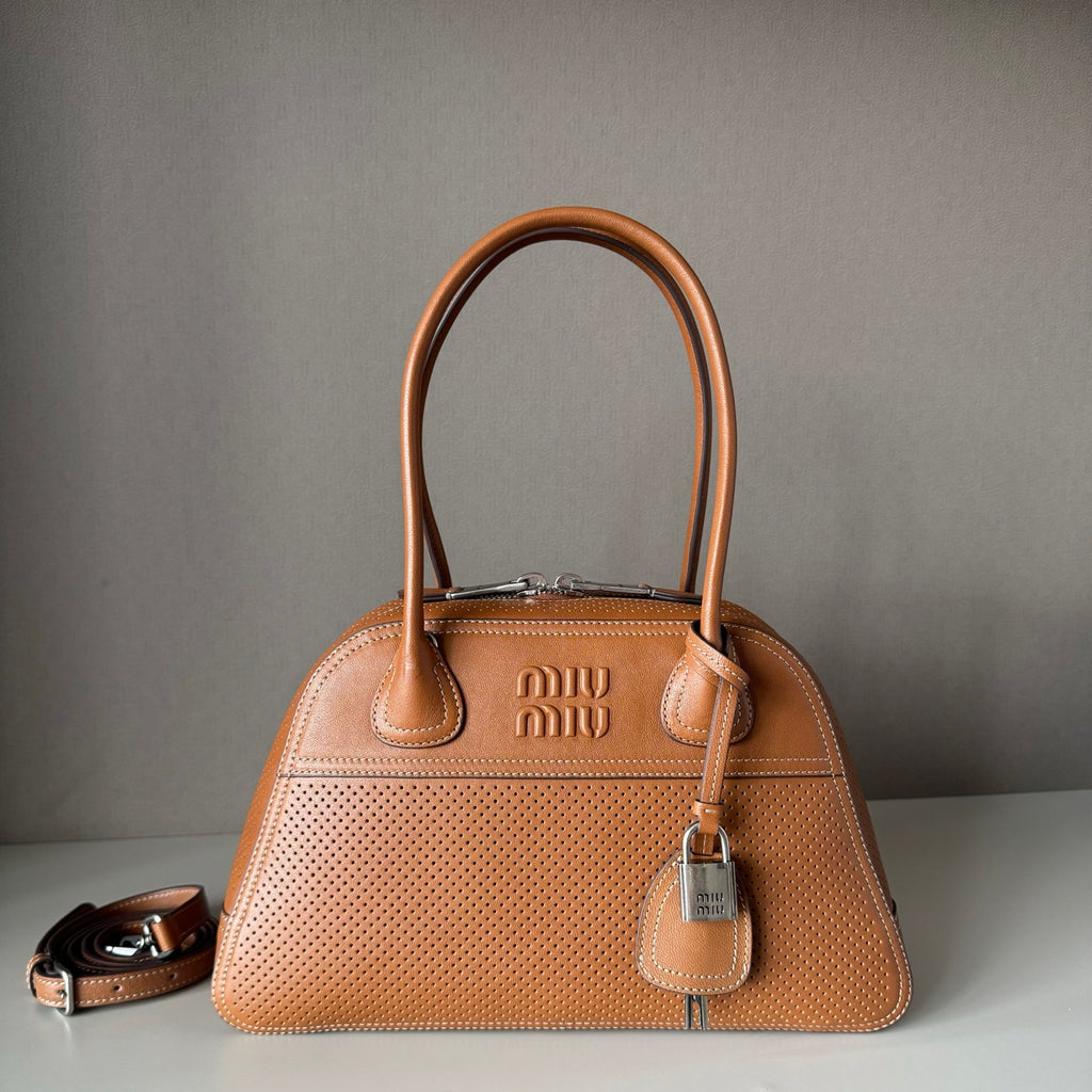 A tan Miu Miu leather handbag with perforated detailing, dual top handles, a hanging padlock, and a detachable shoulder strap, displayed on a light surface against a gray background.